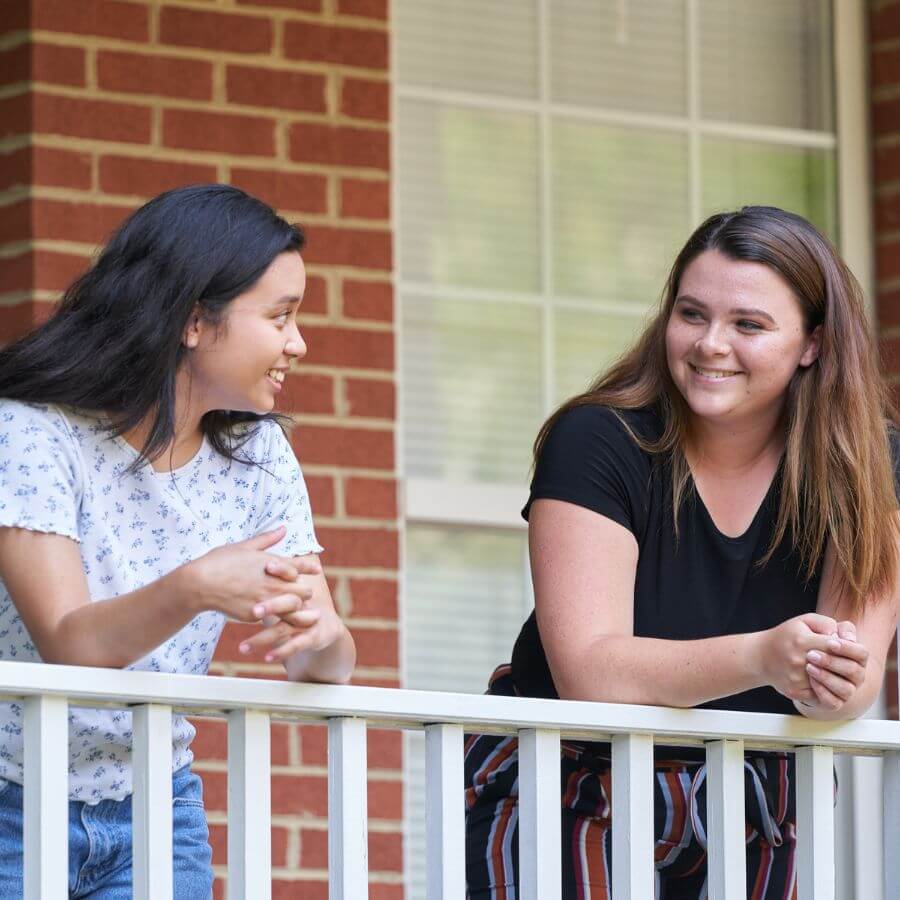 Two teen girls smiling at each other while leaning towards a fence