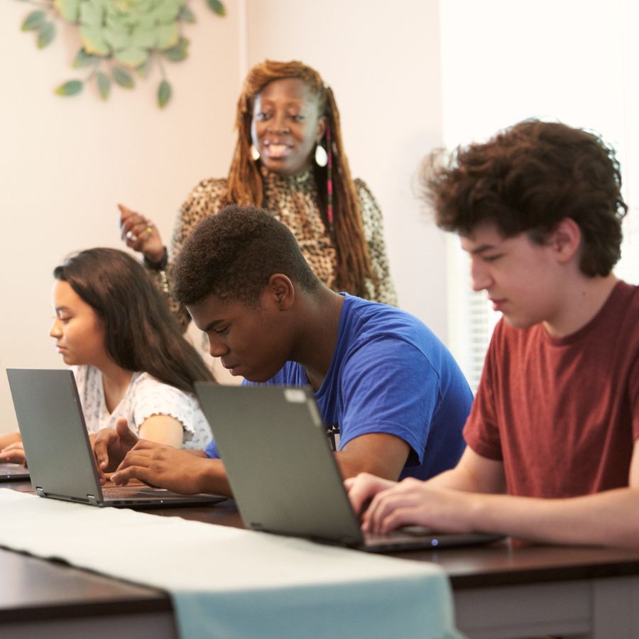 Teens studying with laptops in a big table together and teacher's giving them guides