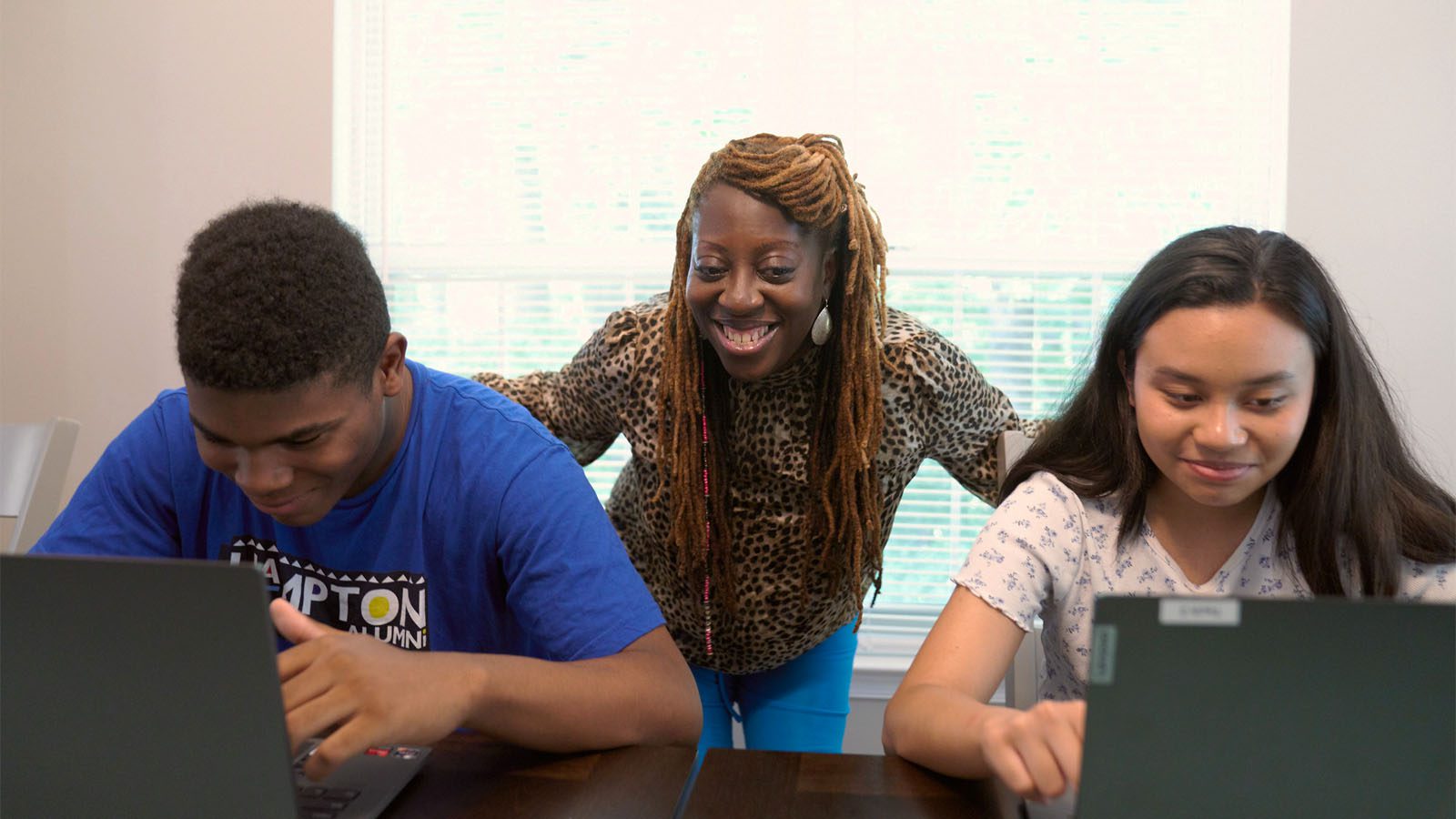 A smiling woman assisting two focused teenagers working on laptops at a table.
