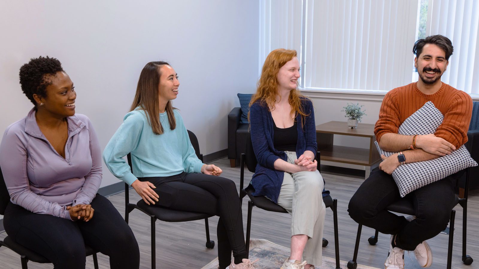 Four young adults smiling and engaged in a conversation while seated in a circle during a group therapy session in a room with soft natural lighting.