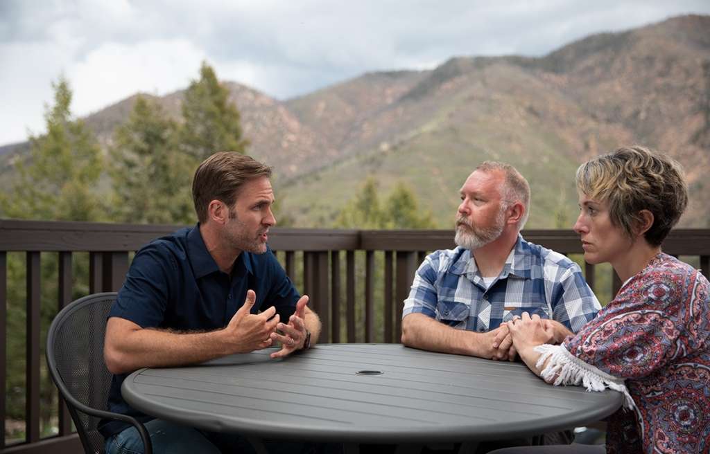 A mom and did listen and hold hands as they sit across from a clinician explaining treatment at a table outdoors.