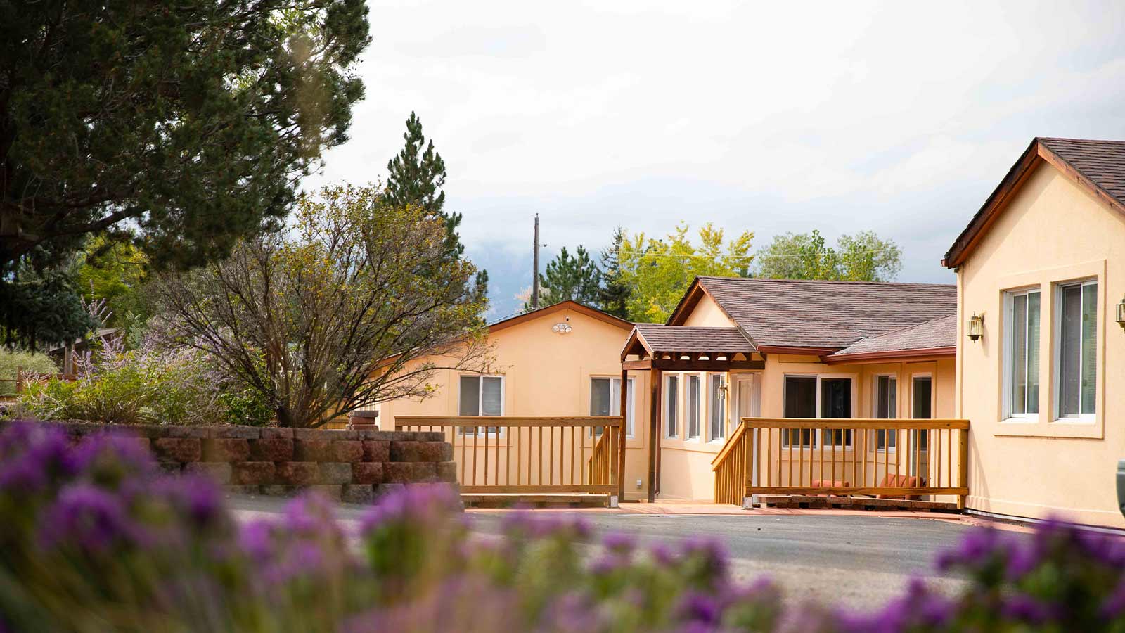 A suburban home with a welcoming wooden front porch, surrounded by lush greenery and blooming purple flowers.