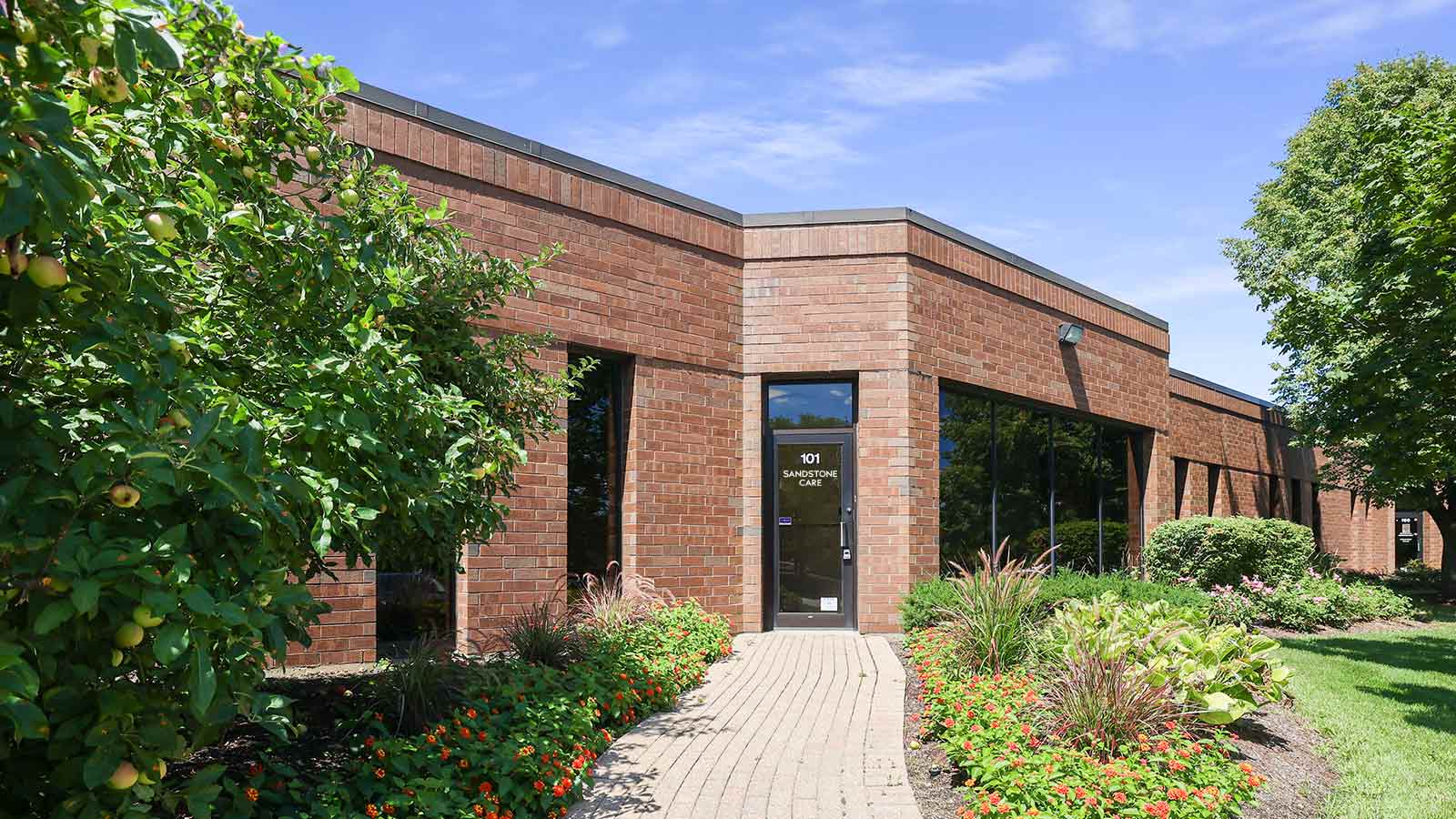 A single-story brick building with a clear entrance pathway lined with flowering shrubs.