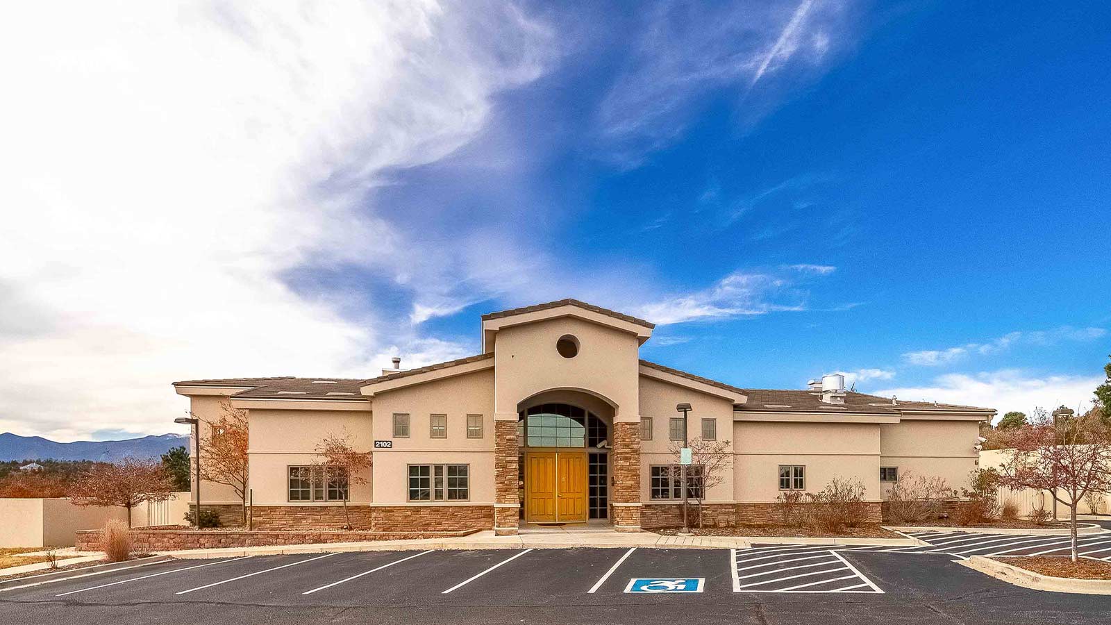 A single-story tan stucco building with a large central entrance and parking in front under a vast blue sky.