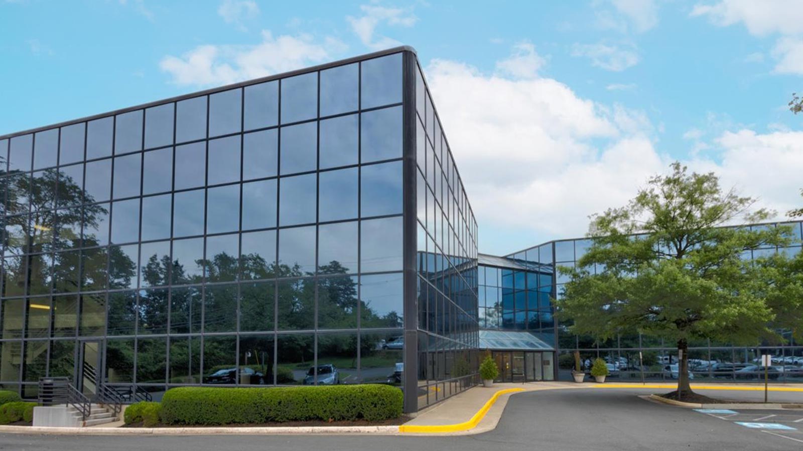 A modern glass building surrounded by trees and a parking lot under a blue sky.