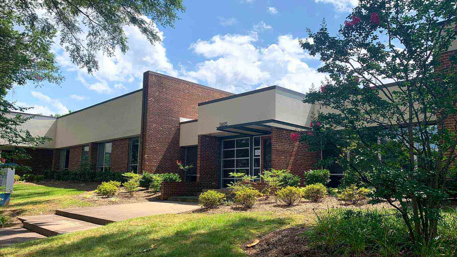 A brick office building with a covered entrance, surrounded by greenery under a blue sky.