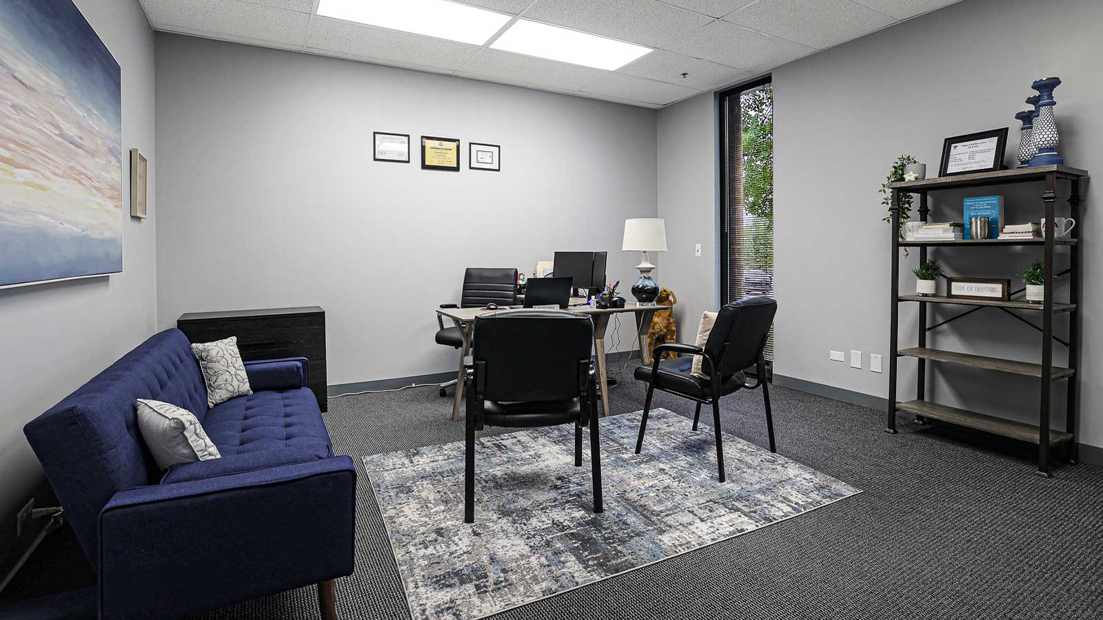 An office space with a blue sofa, desk, chairs, bookshelf, and framed certificates on the wall.