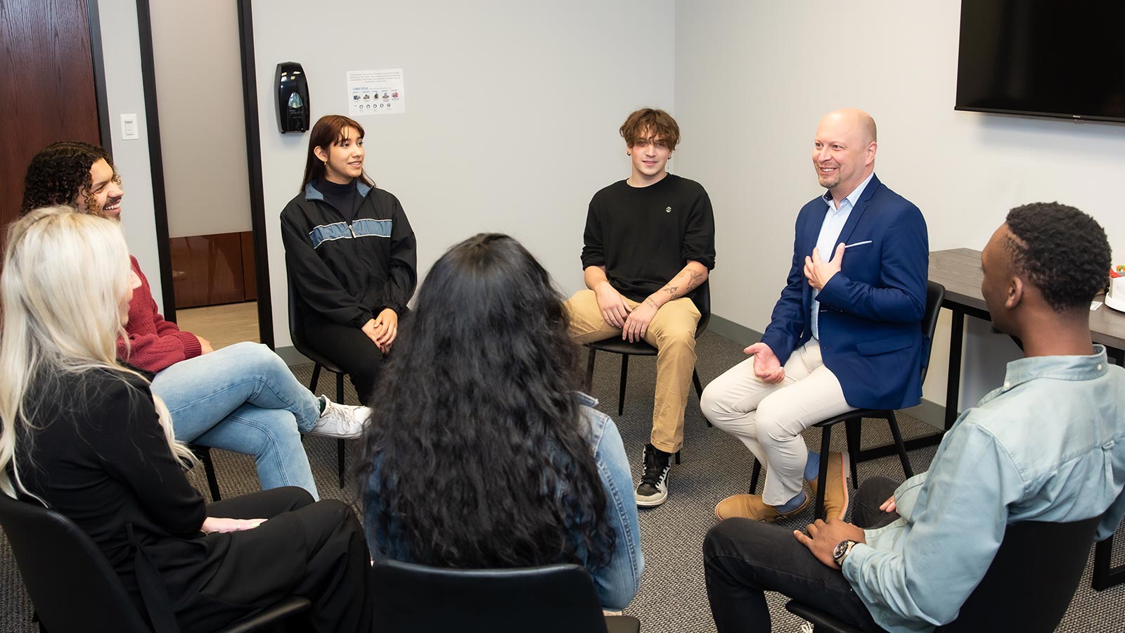 A group therapy session with a facilitator speaking to young adults in a circle.