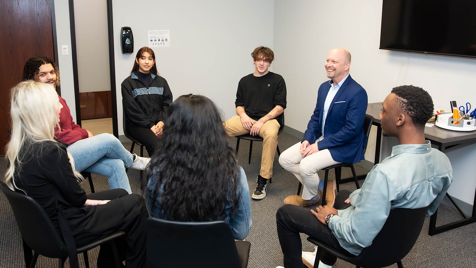 A group of young adults seated in a circle during a group session, smiling and engaging with a counselor.