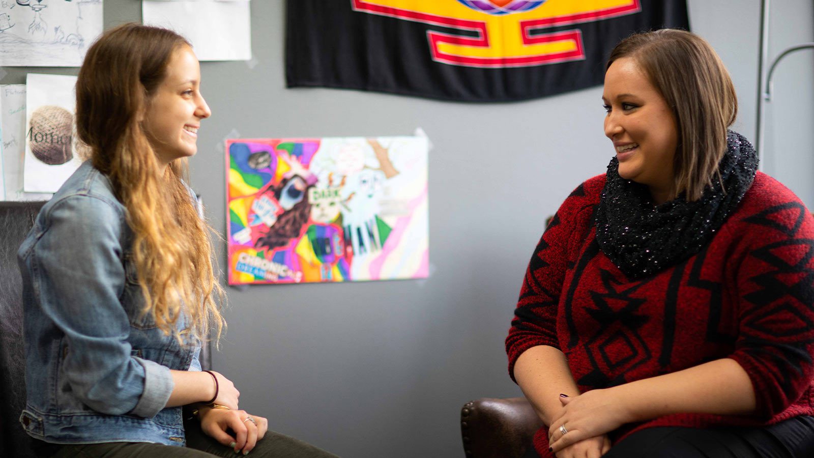 Two women having a cheerful conversation in an office with colorful posters on the wall.