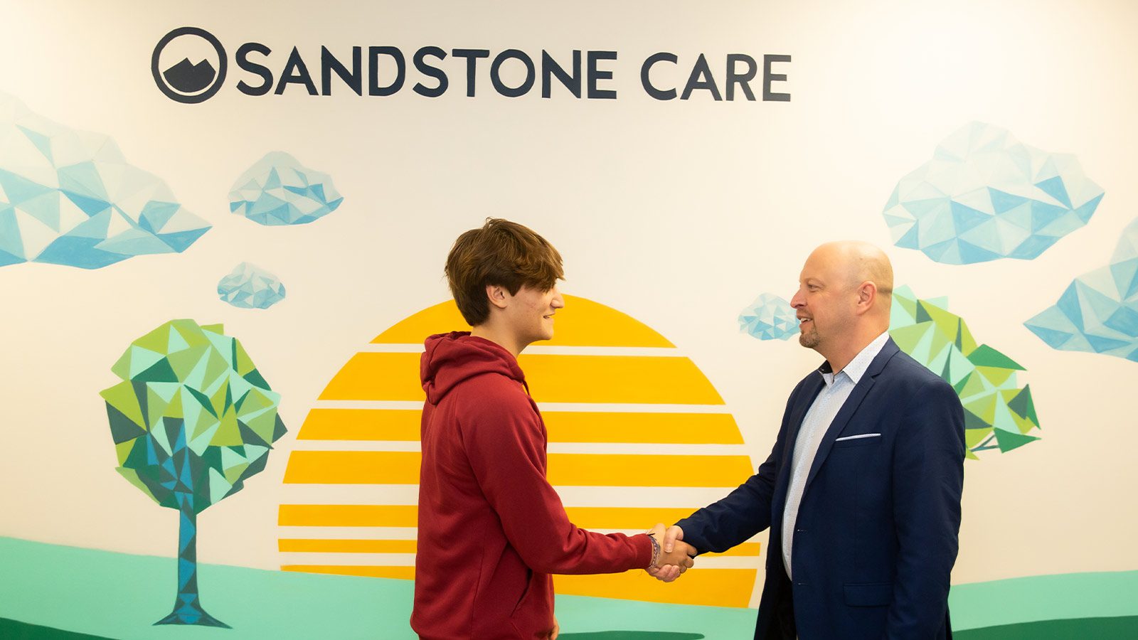 A teenager and man shaking hands in front of a mural with the logo "Sandstone Care."