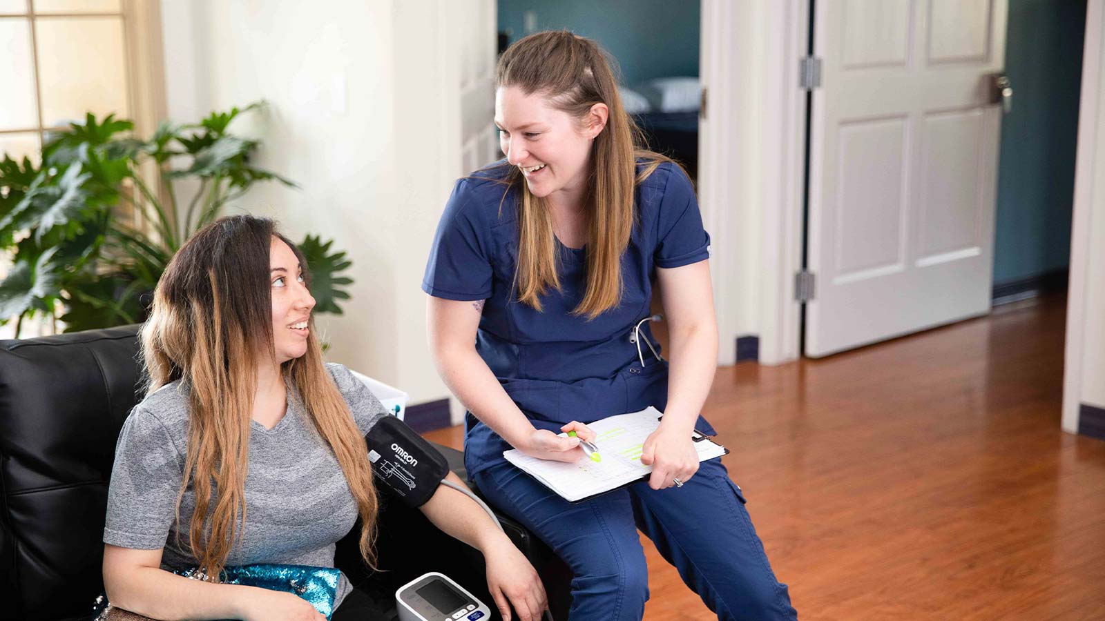 A nurse in blue scrubs is sitting and taking notes while talking to a seated woman in a casual setting.