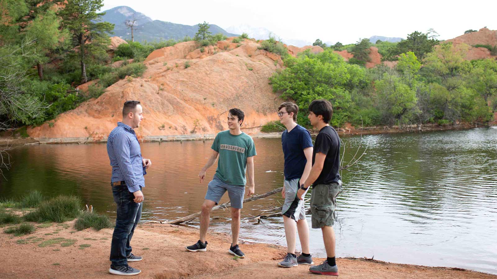 Four individuals standing by a lakeside, surrounded by natural red rock formations and greenery.