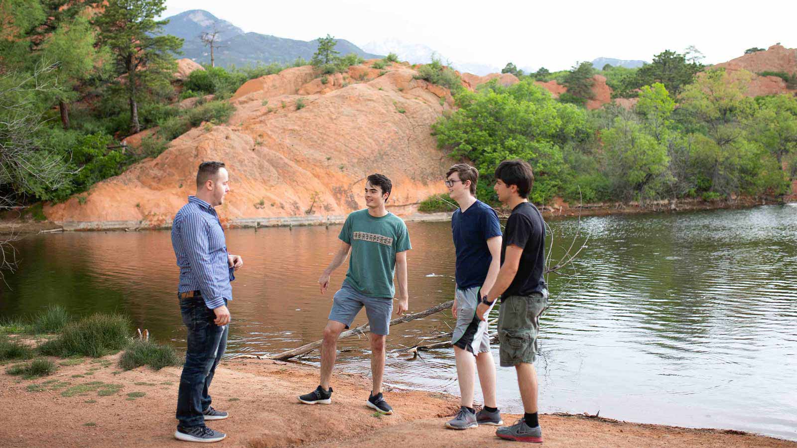 Four individuals standing by a lakeside, surrounded by natural red rock formations and greenery.