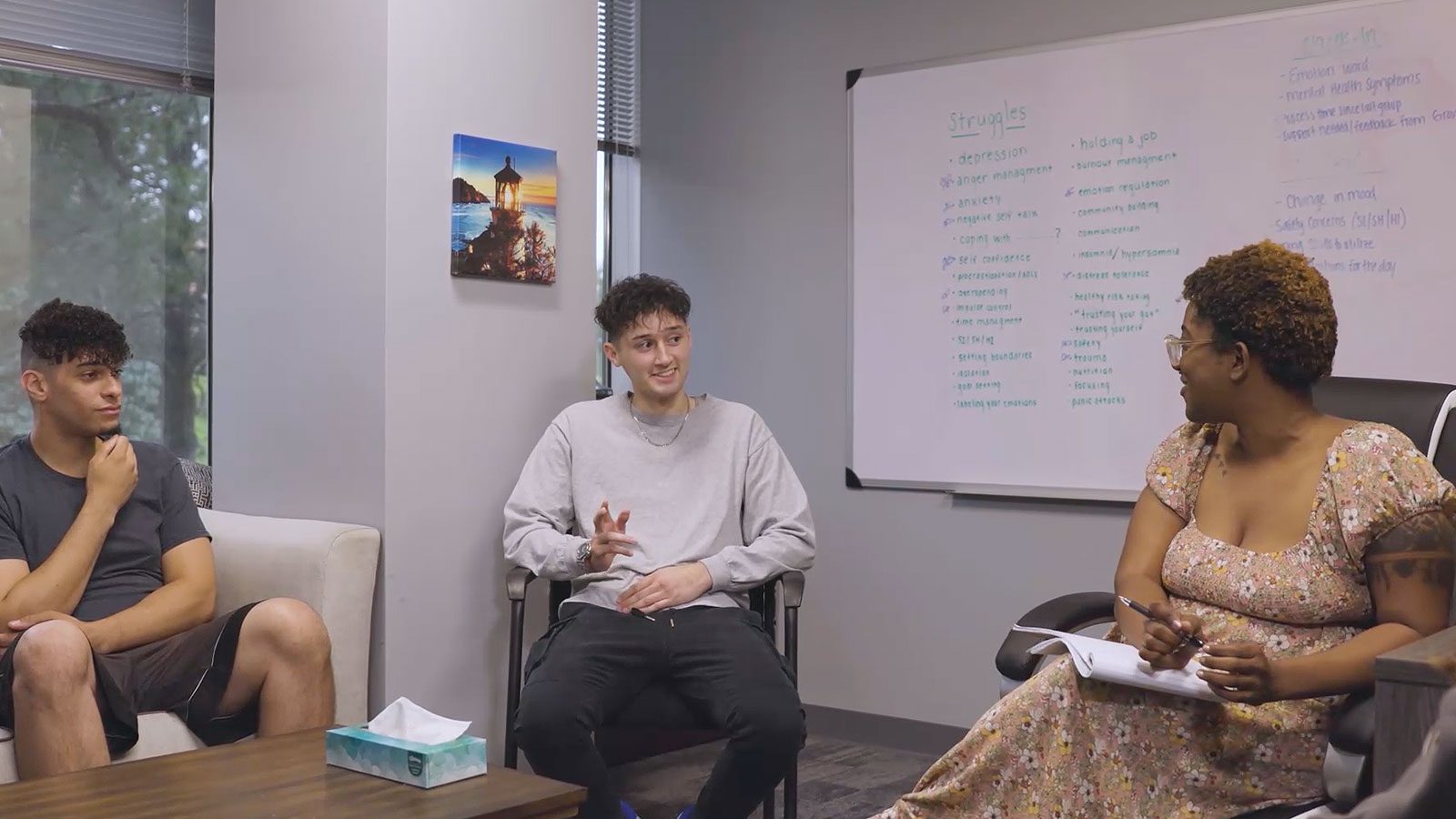 A group therapy session with young adults and a facilitator discussing topics written on a whiteboard.