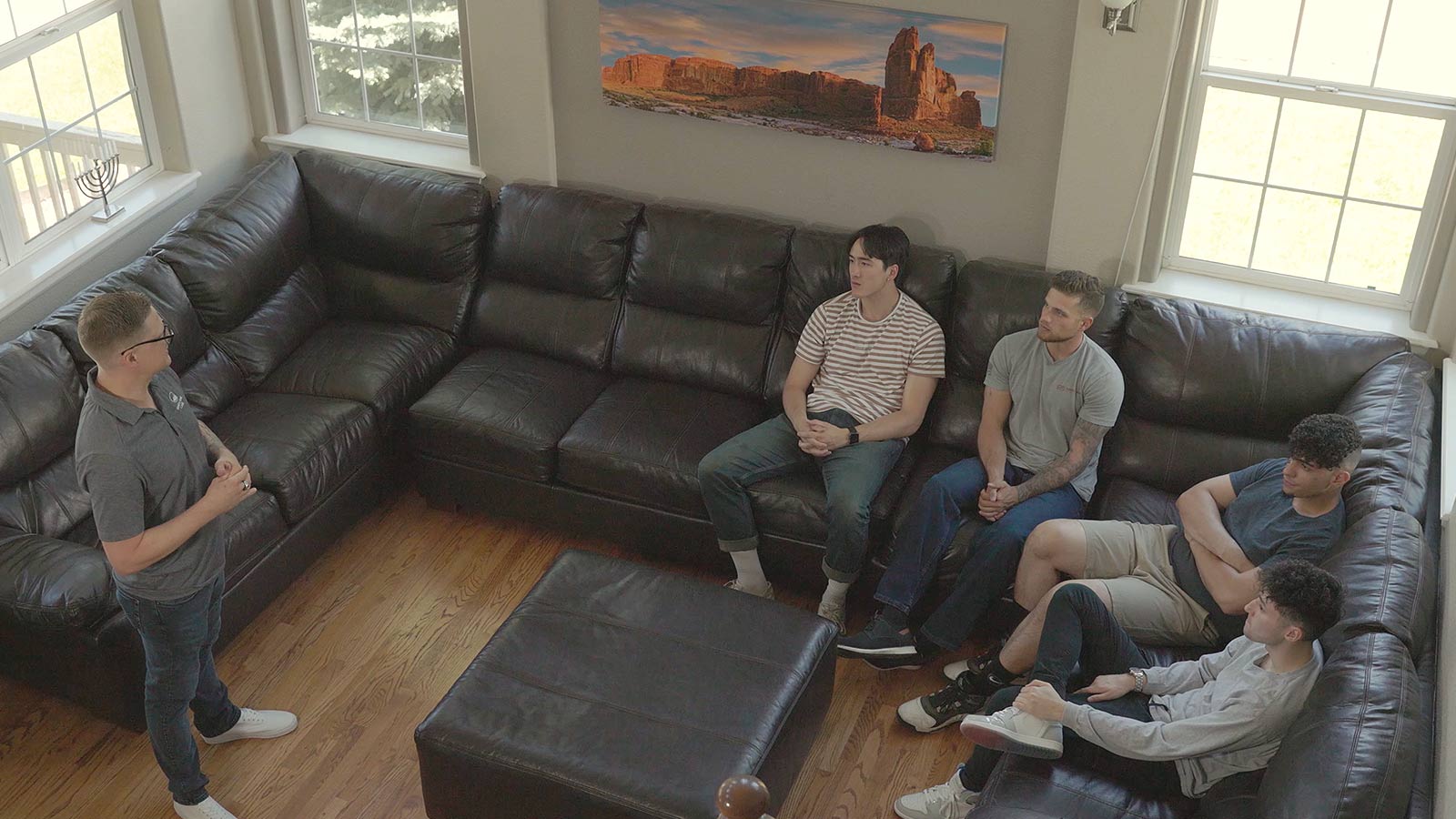 Four men listening to another man speaking in a living room with a large sectional sofa.