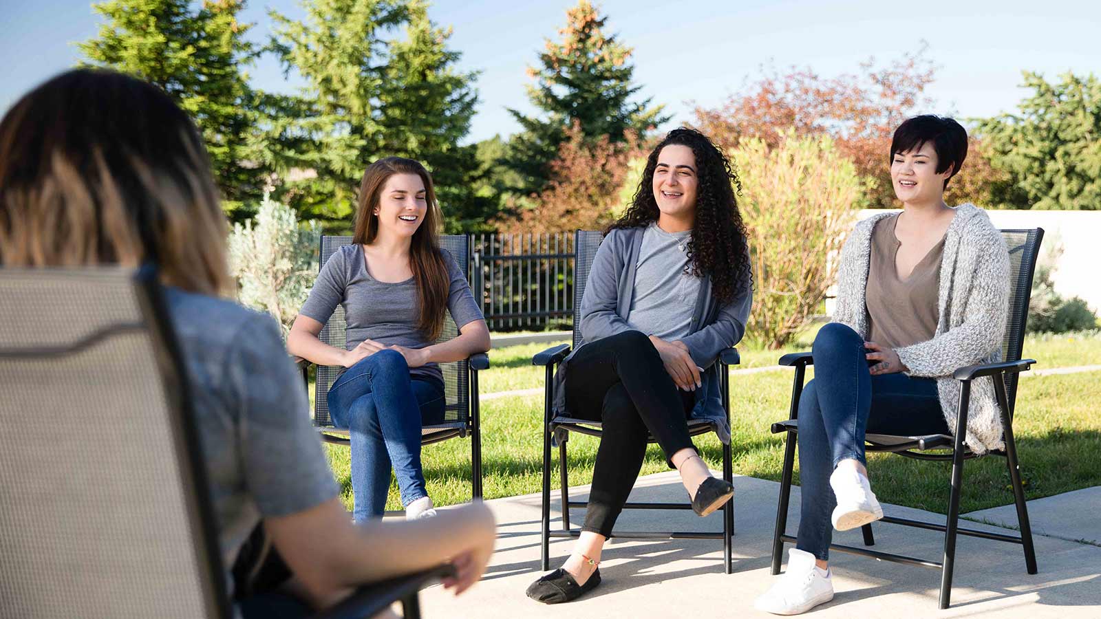 Four women are engaged in conversation while sitting in a circle of chairs outdoors.