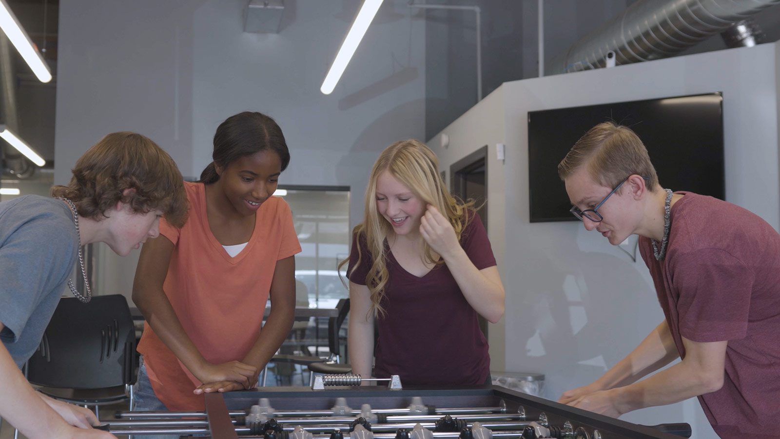 Teenagers smiling and playing foosball in a bright, contemporary recreational space.