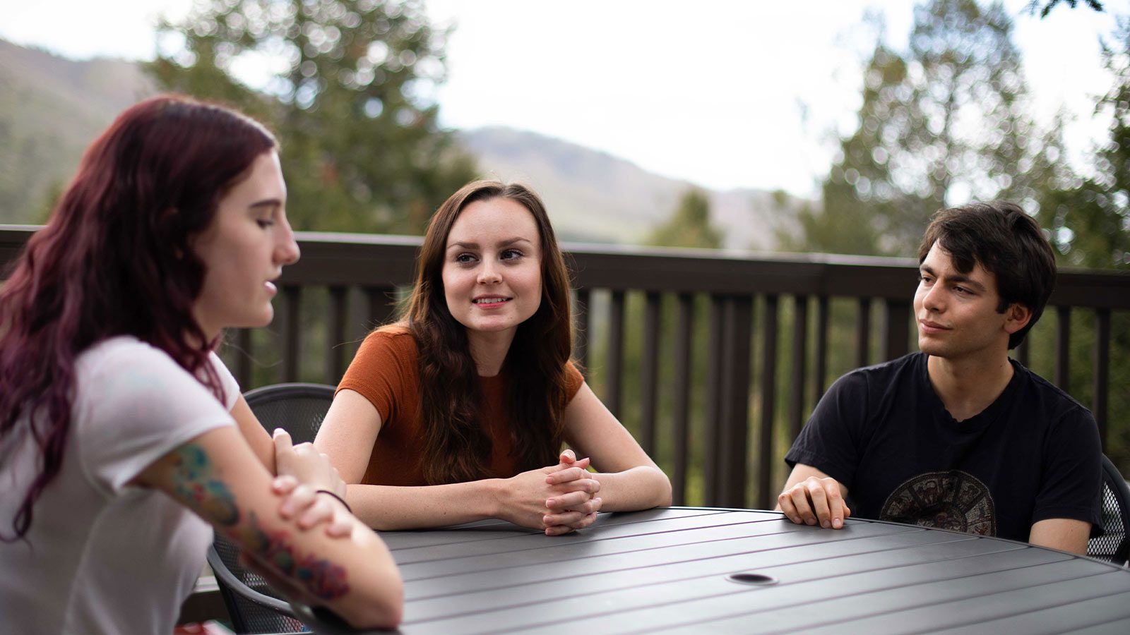 Three young adults engaged in a conversation around a patio table with a backdrop of trees and hills.