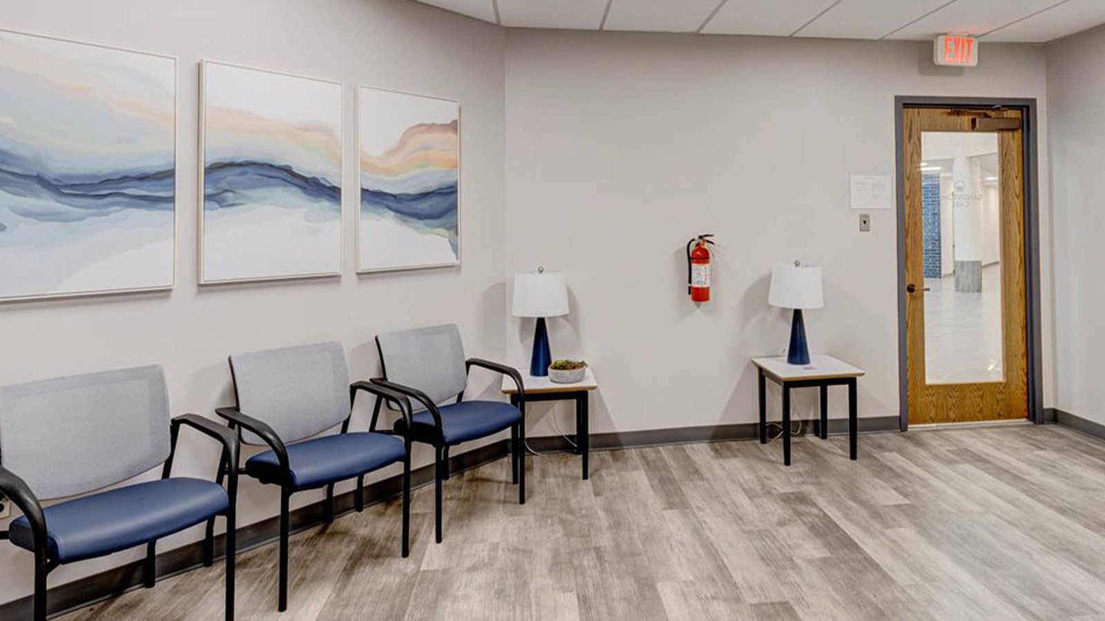 A waiting area with chairs lined up, artwork on the wall, and a side table with a lamp.