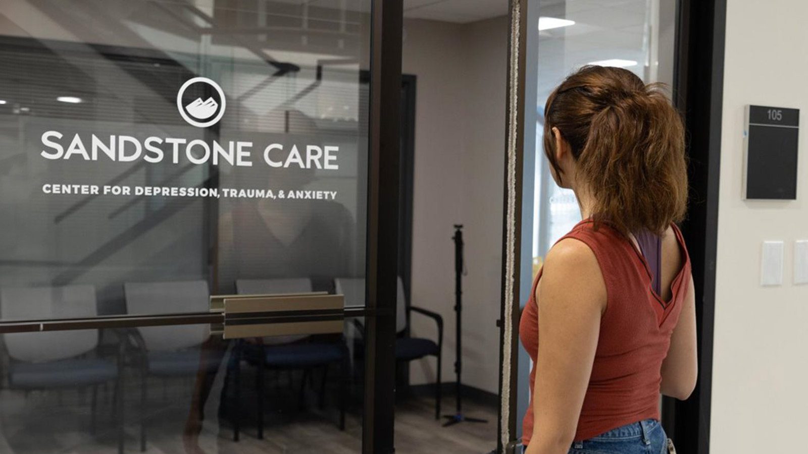 A woman walking into an office with a sign that reads "Sandstone Care, Center for Depression, Trauma, & Anxiety".