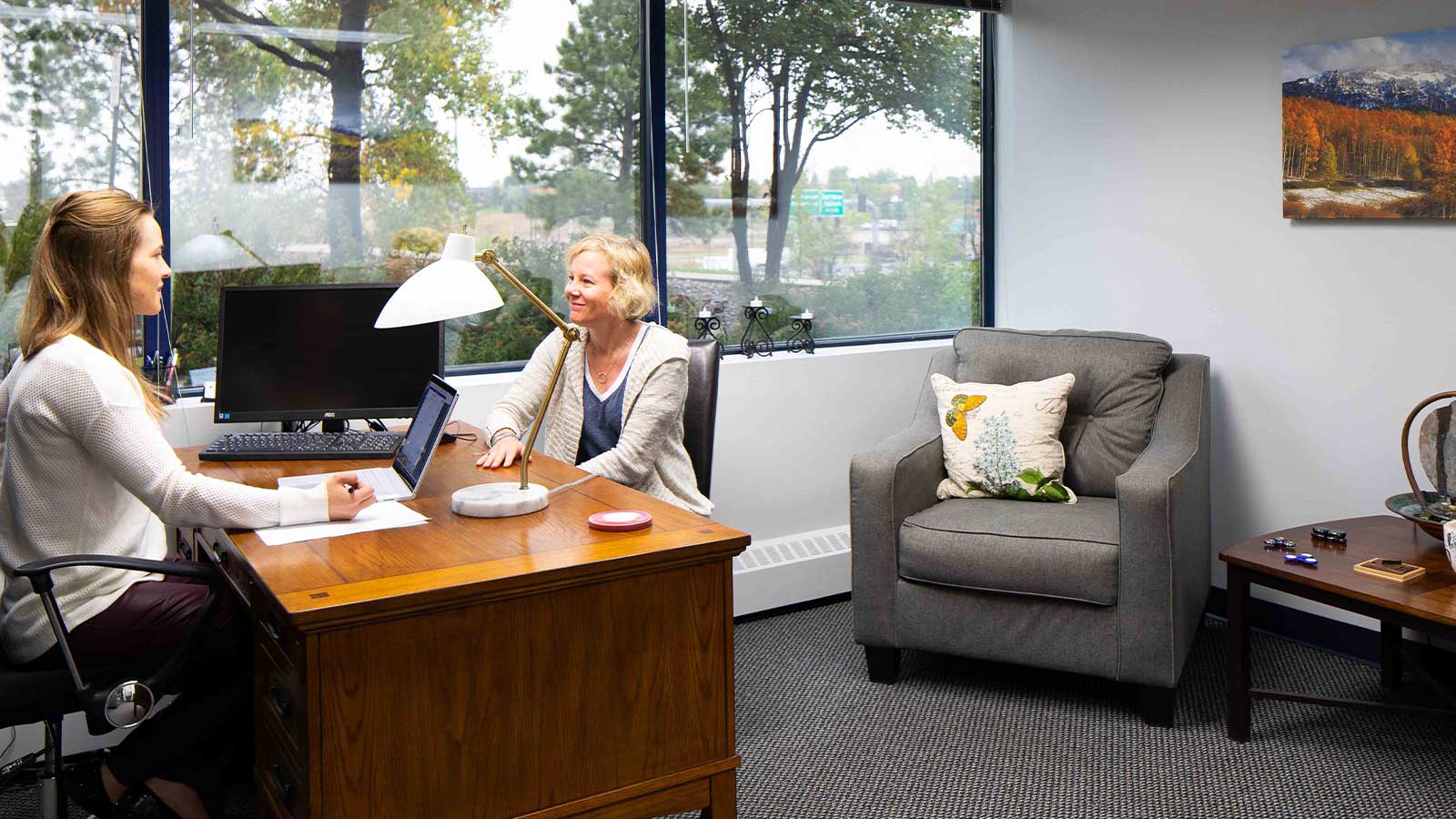 Two women talking in an office space with a wooden desk, computer, comfortable chairs, and nature views.