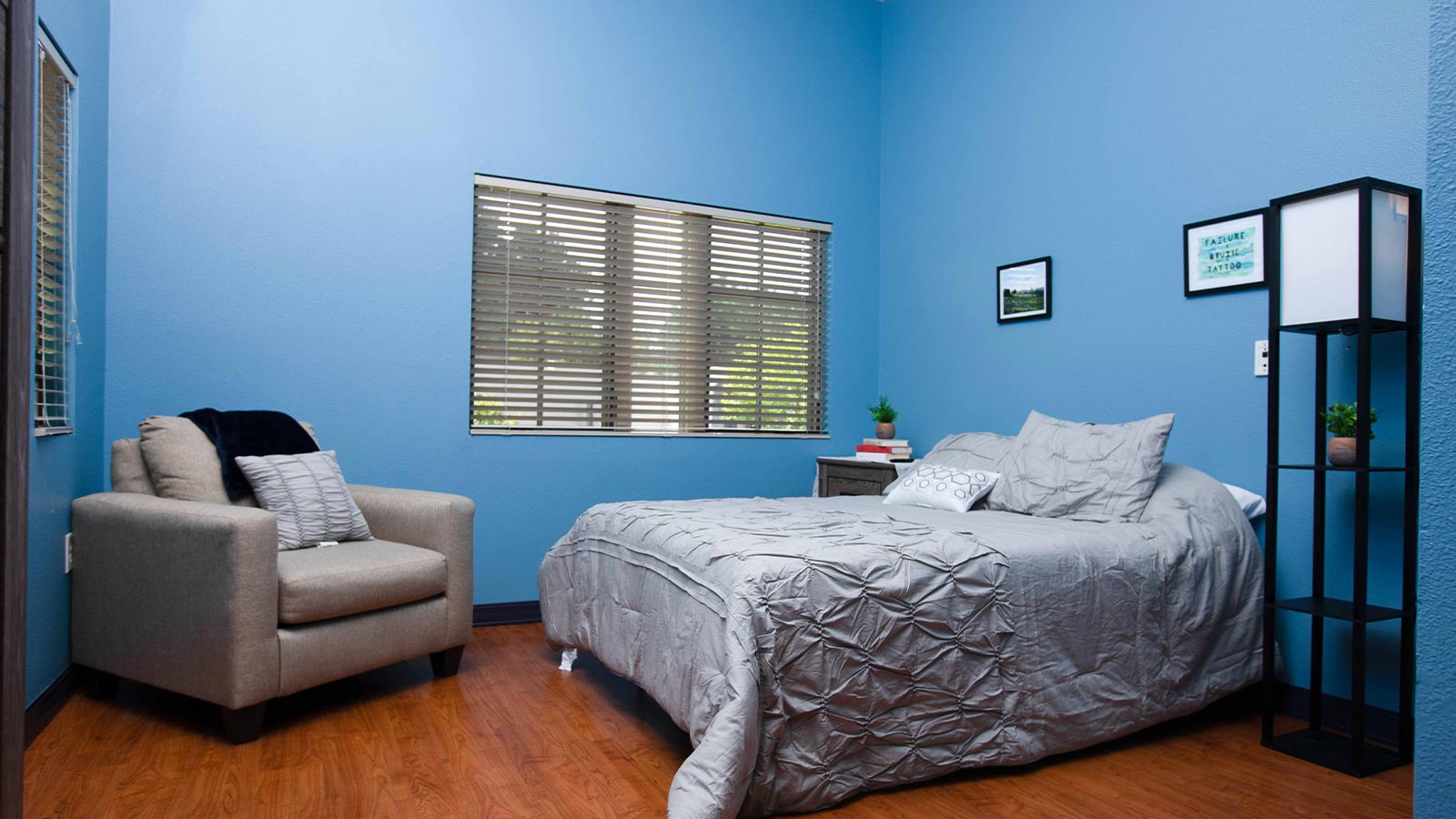 A minimalist bedroom with a blue wall, a bed with a gray duvet, a small sofa, and a floor lamp.