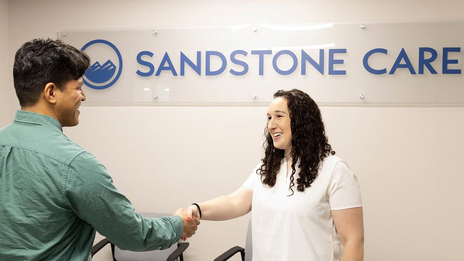 Two people smiling and shaking hands in an office with a blue Sandstone Care logo in the background.