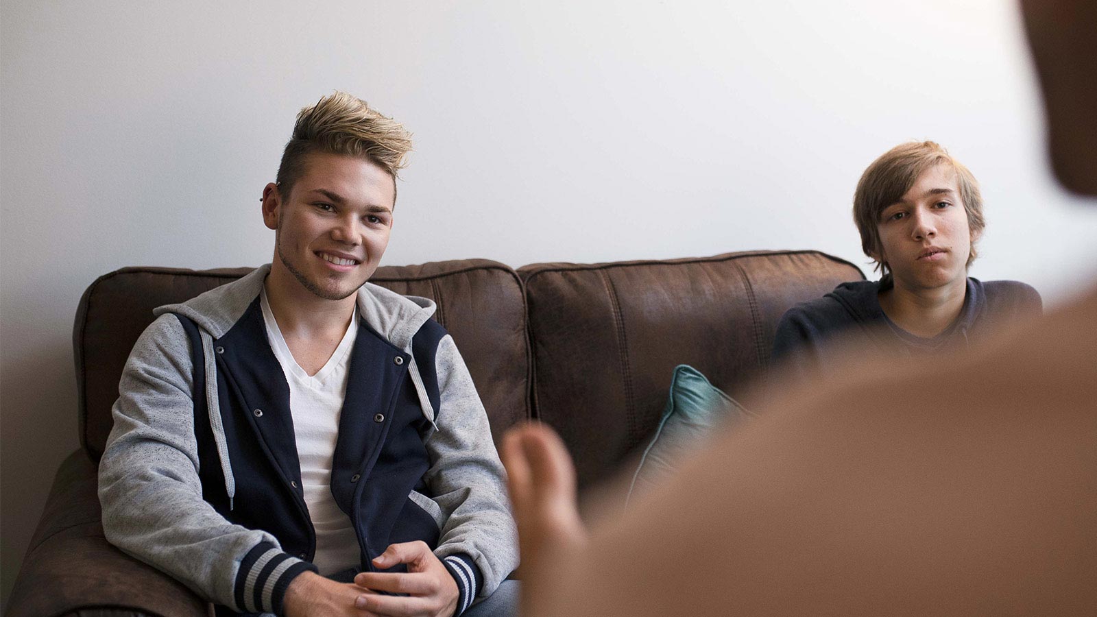 Two individuals seated on a couch in a group therapy session, one speaking and the other listening attentively.