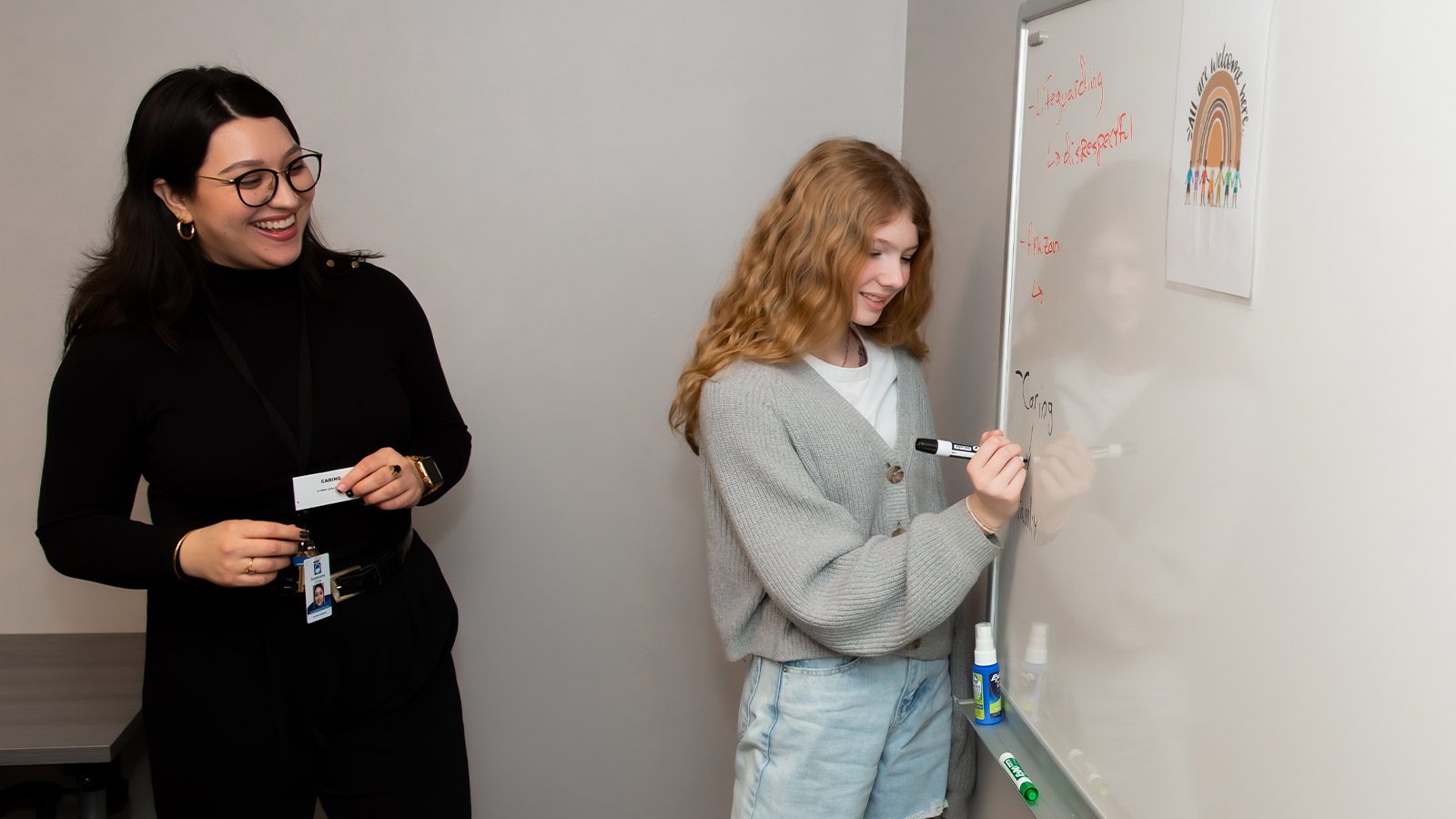 A young woman writing on a whiteboard in a well-lit room with the teacher observing.