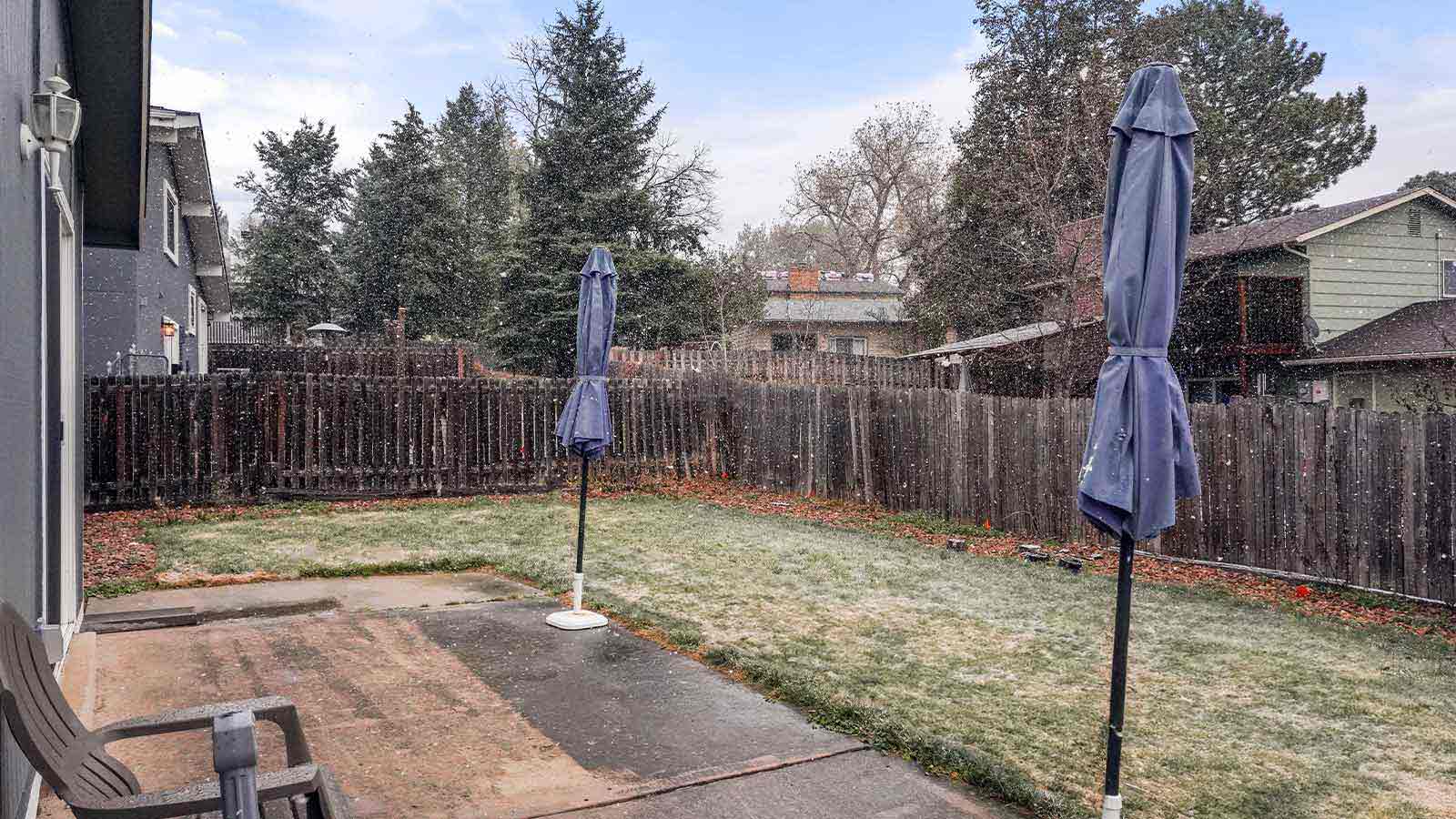 A backyard under snowfall, with a patio and two folded umbrellas.