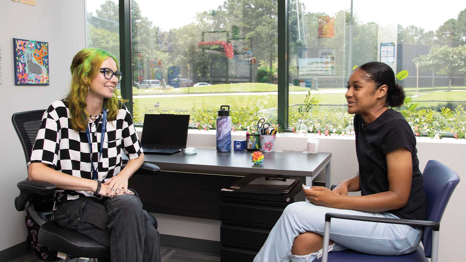 there are two women sitting at a desk talking to each other