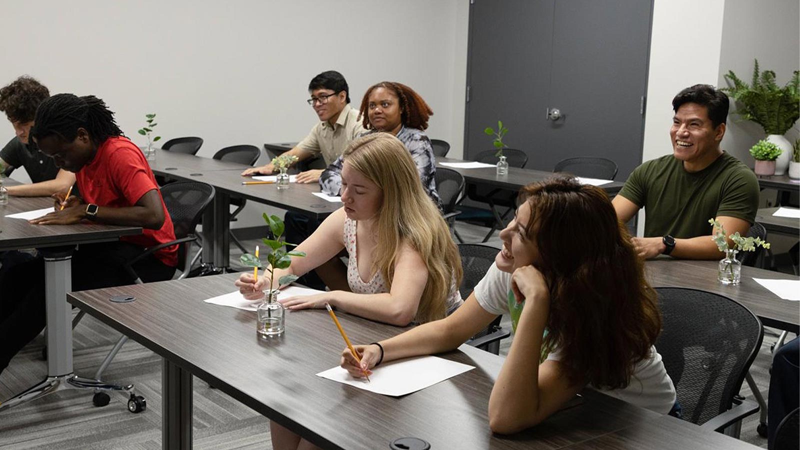 A diverse group of seven people in a classroom, some taking notes and some interacting with each other.