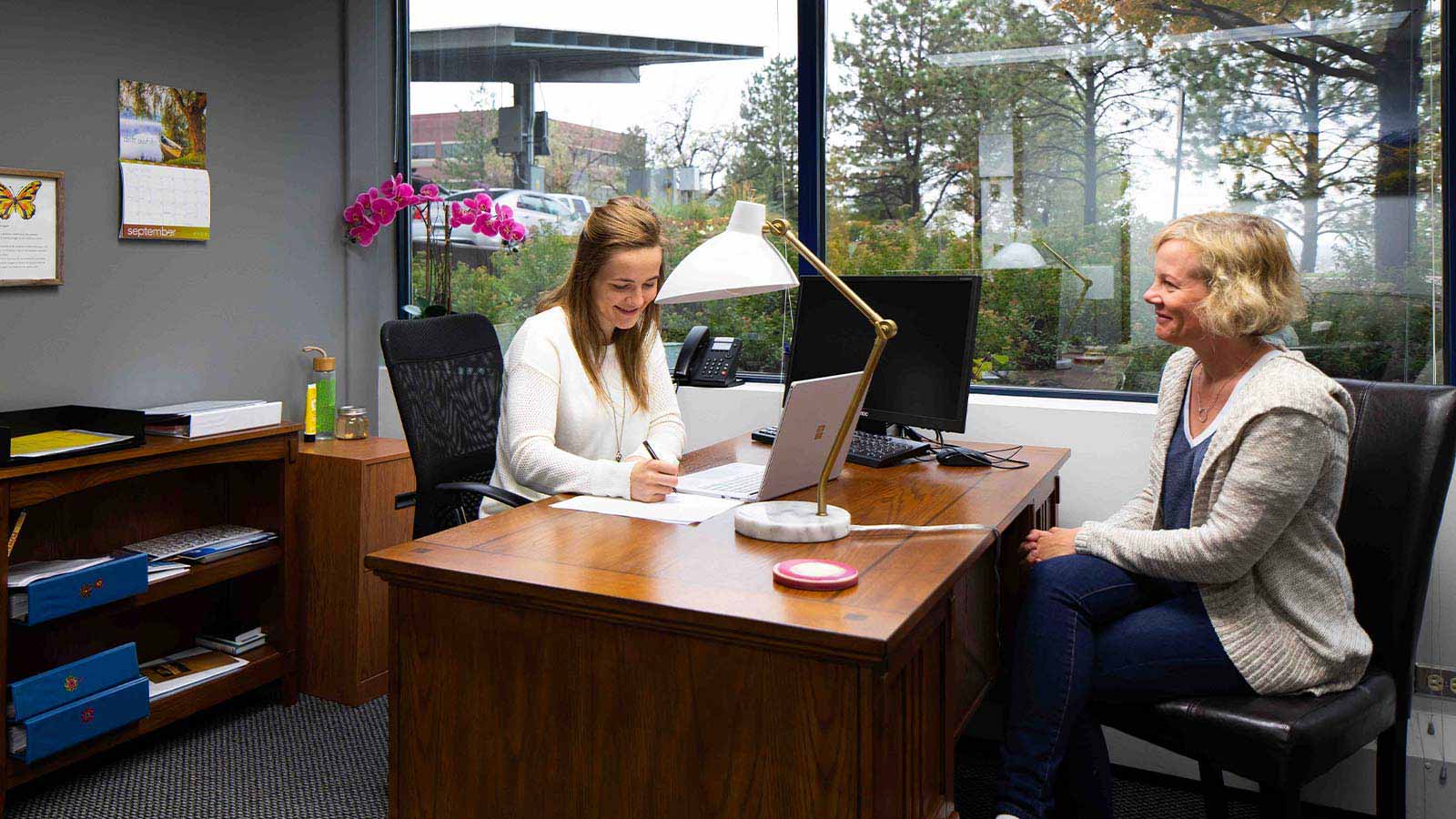 A professional smiling at a woman sitting across from her in an office with a view of the outdoors.