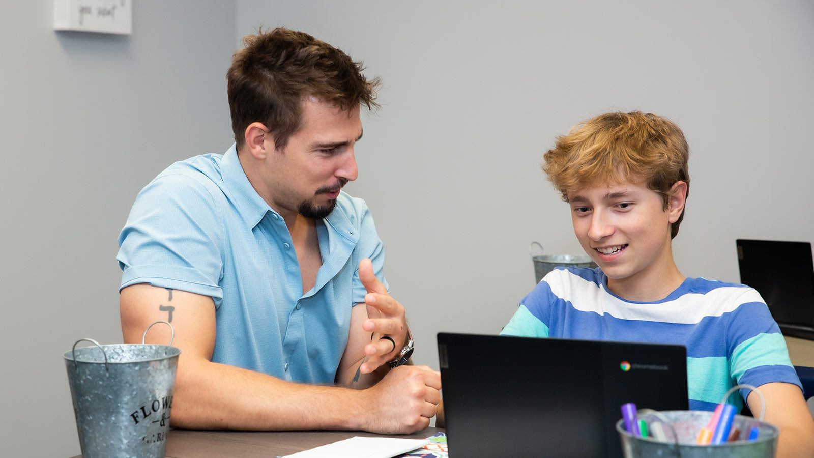 A man and teenage boy engaging in a one-on-one conversation across a desk with a laptop.