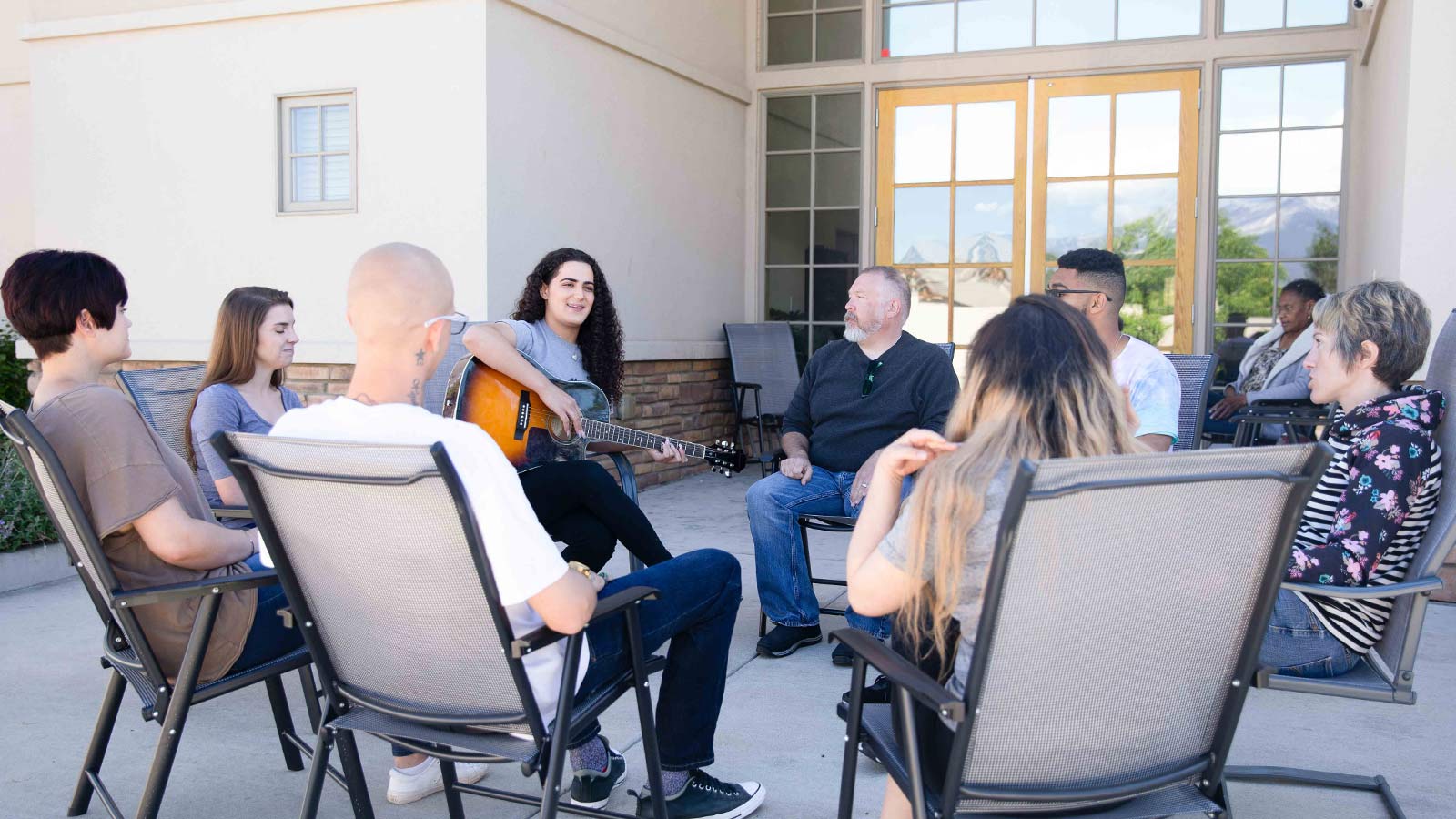 A group of people seated in chairs outdoors in a circle with a woman playing guitar and singing.