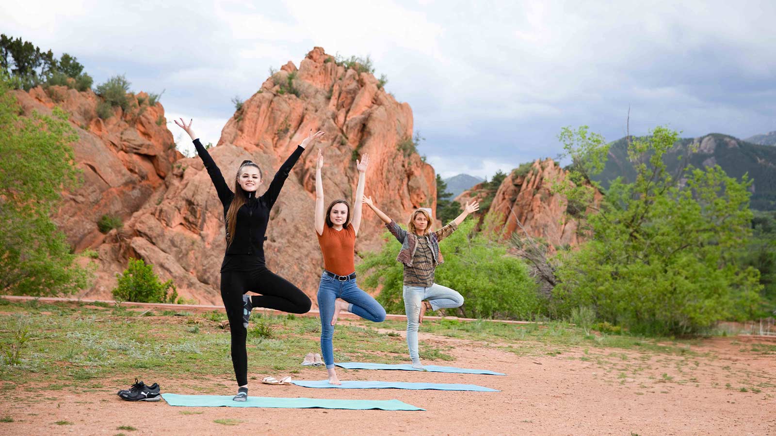Three individuals performing yoga poses on mats in an outdoor setting with red rocks and greenery in the background.