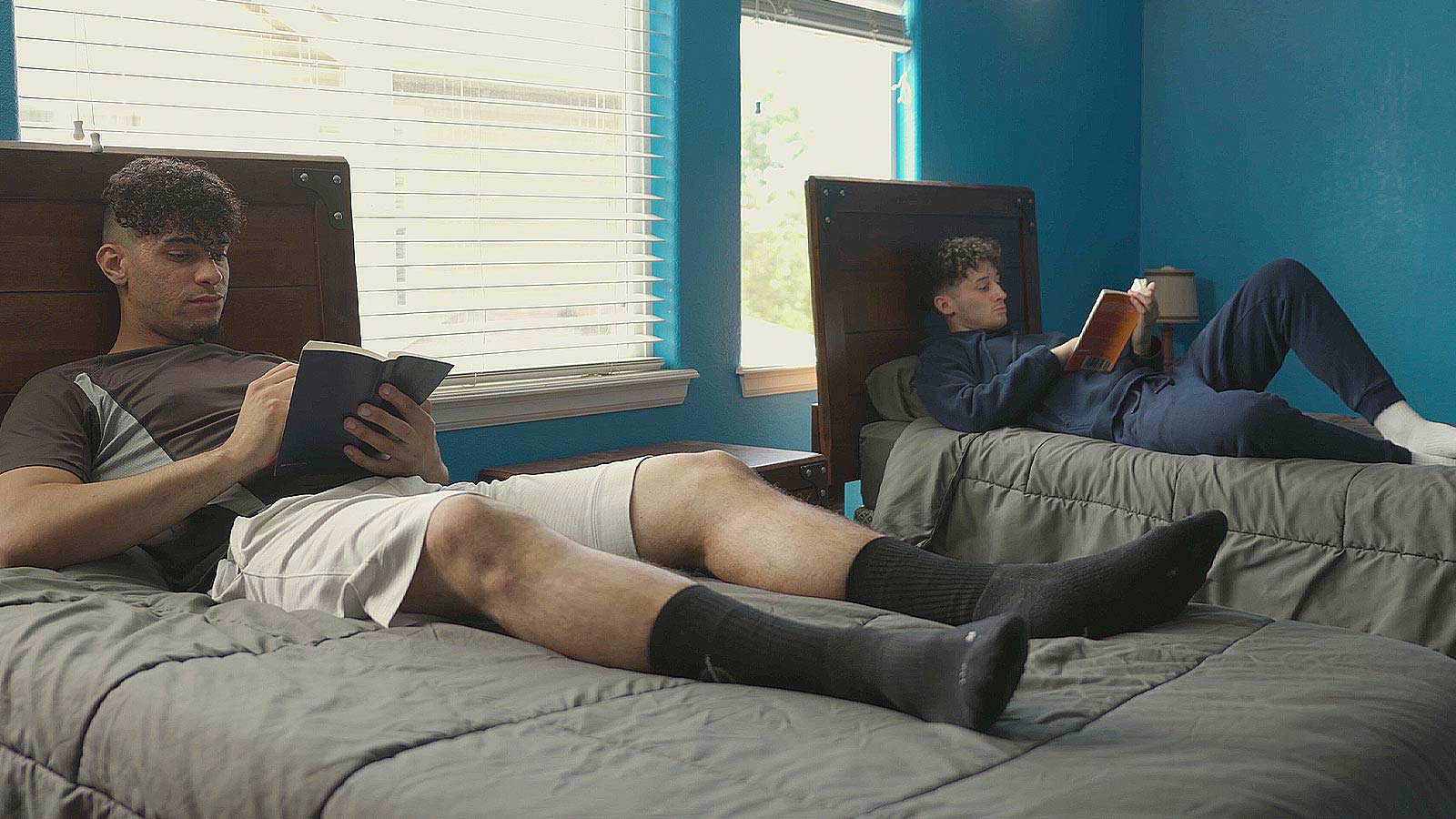 Two men reading books while relaxing on beds.