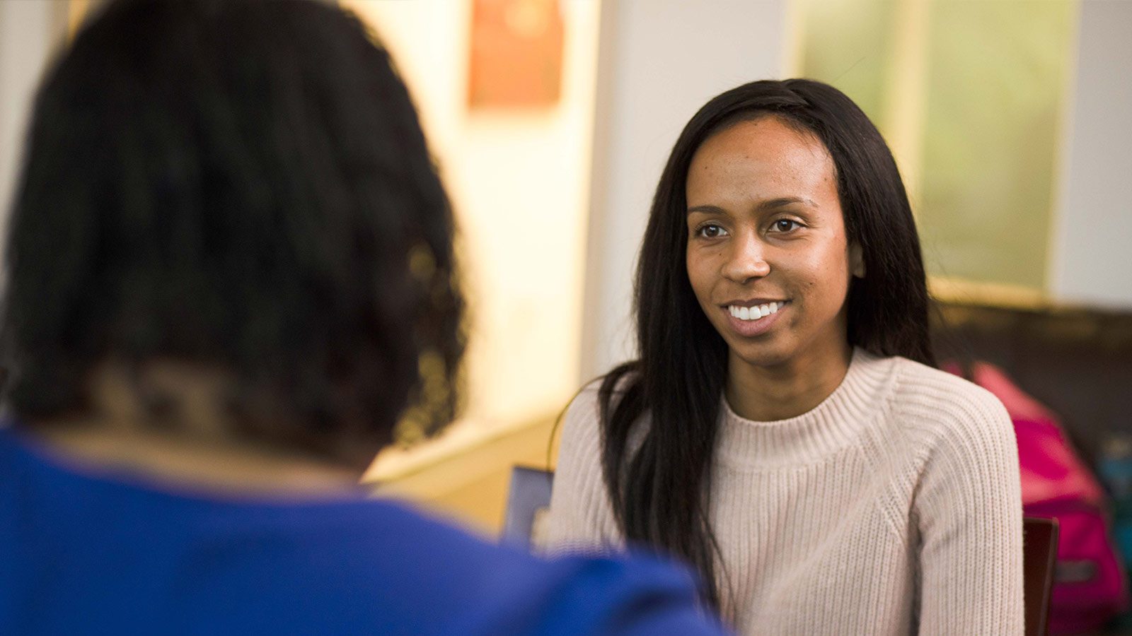 A smiling woman having a conversation with a person whose back is to the camera in a room with soft lighting.