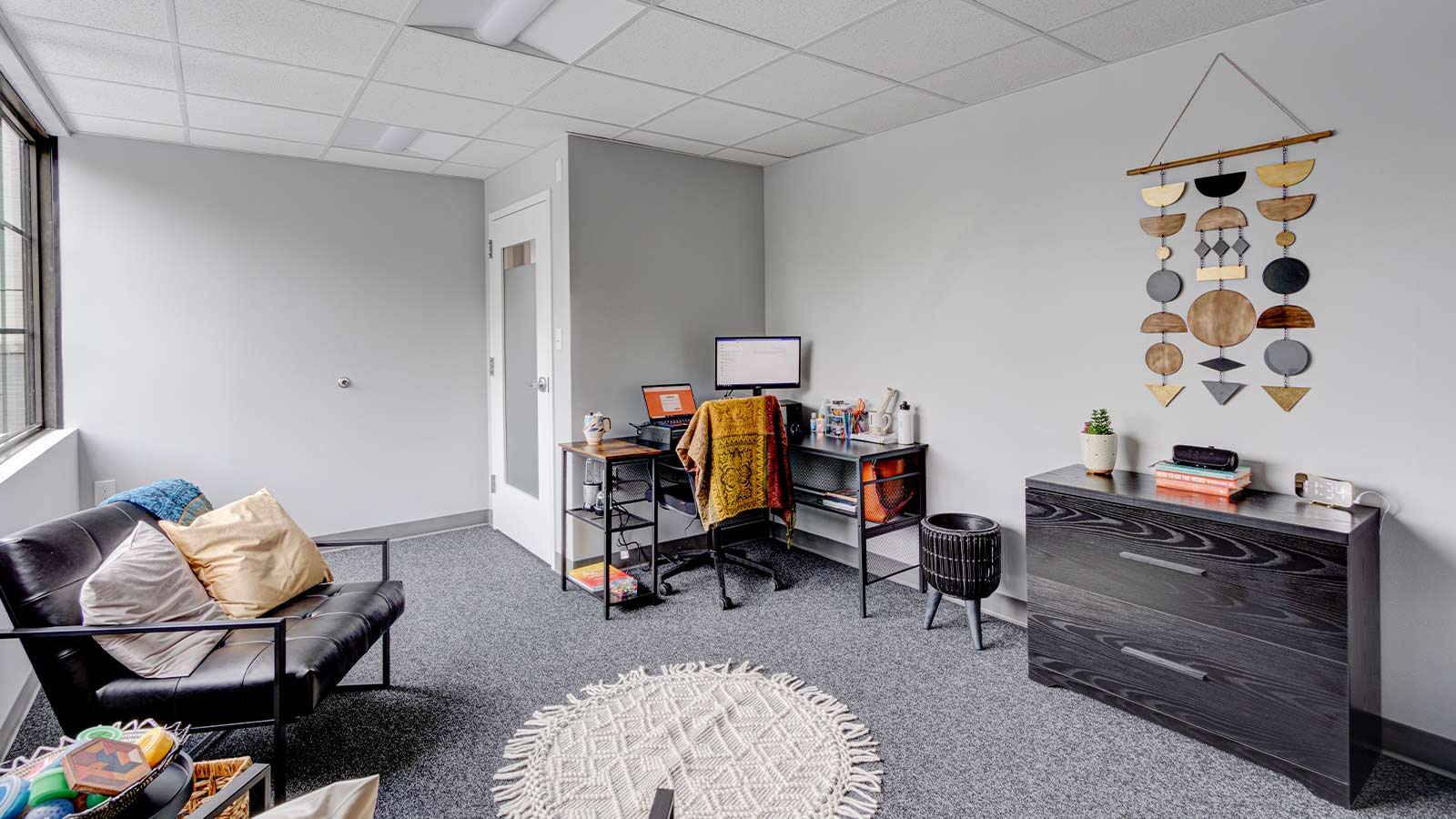 A cozy and modern office space featuring a black sofa, a desk with a computer, and a decorative wall hanging.