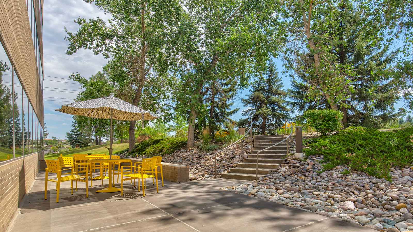 An outdoor seating area with a patterned umbrella and bright yellow chairs, next to a staircase leading to a wooded area.