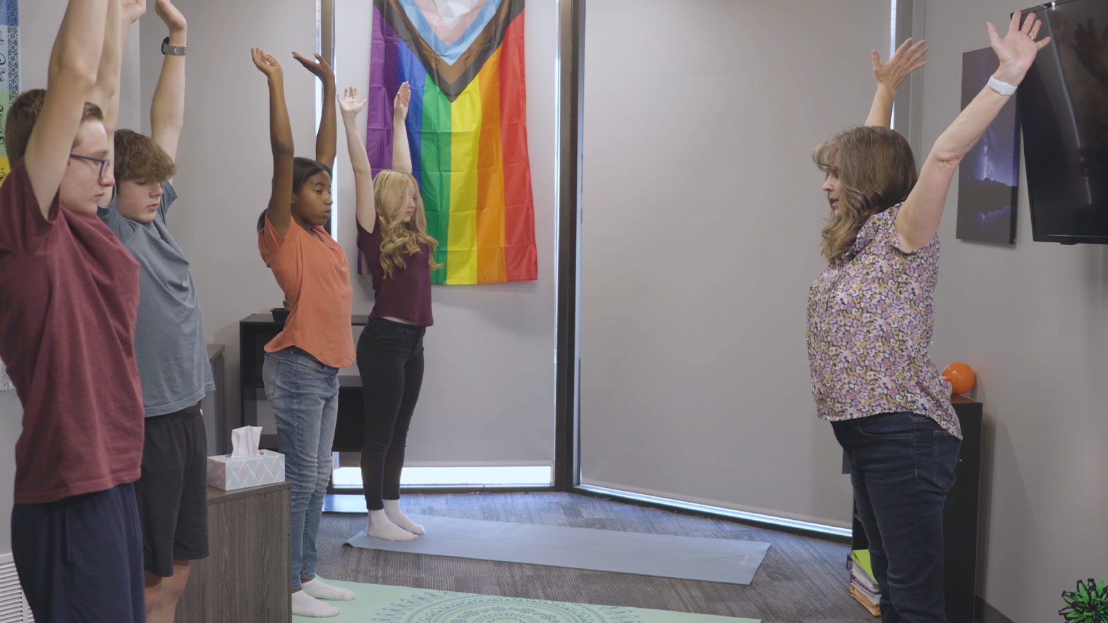 Participants stretching their arms up during a group physical wellness activity in a room with a pride flag.