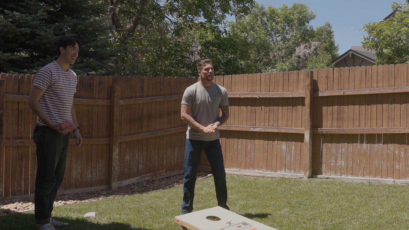 Two men playing a bean bag toss game in a sunny backyard with a wooden fence.