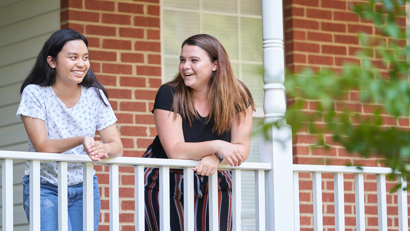 Two young women leaning on a porch railing, smiling and conversing.
