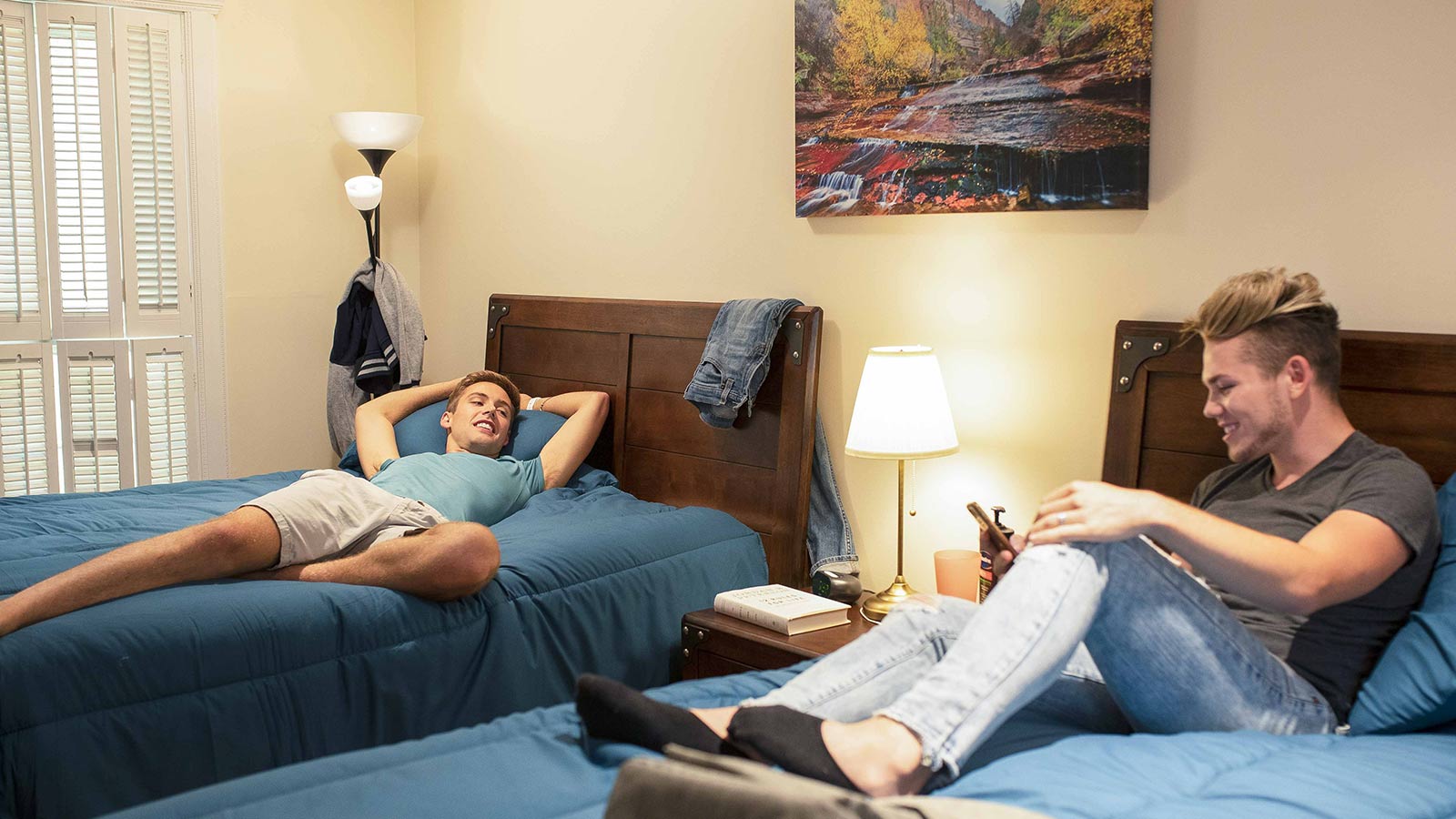 Two men lying on separate beds in a bedroom, one with a phone and the other relaxing.