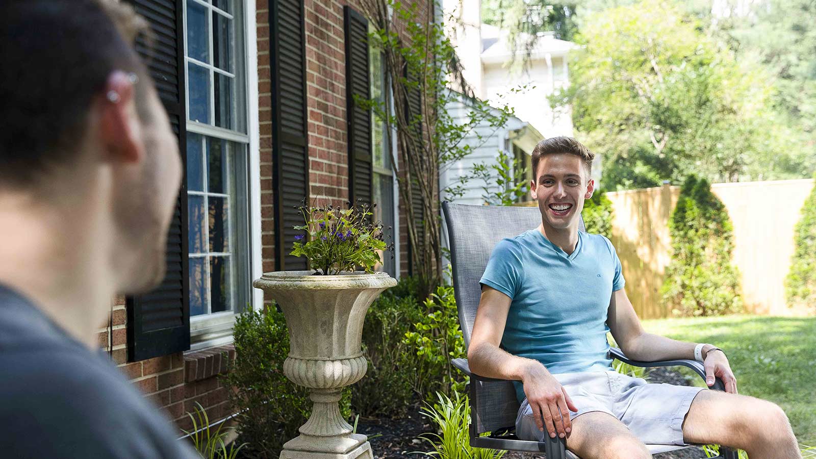 Two individuals relaxing outside a brick home, one smiling and seated comfortably on a patio chair.