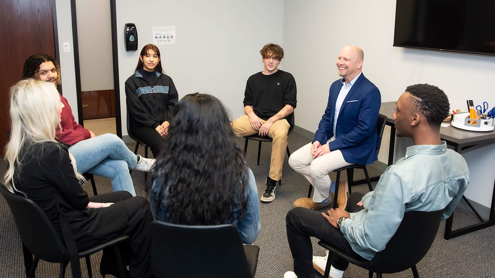 A group of young adults seated in a circle during a group session, smiling and engaging with a counselor.