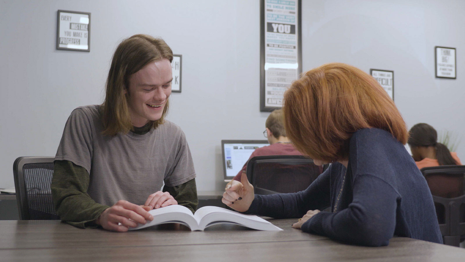 there is a woman sitting at a table with a book and a man sitting at a desk