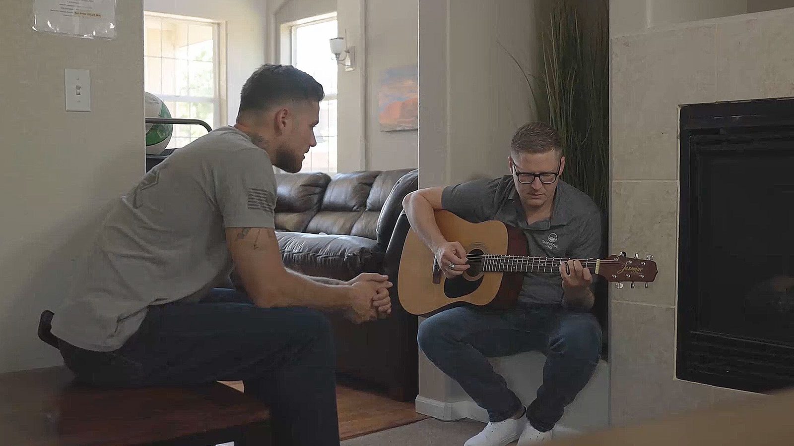 An individual playing an acoustic guitar while another person listens.