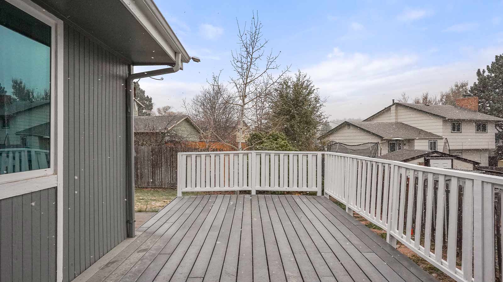 A wooden deck with a railing, overlooking a snowy backyard and neighboring houses.